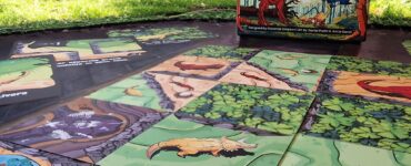 Colorful game board of "Chomp," featuring illustrated dinosaur-themed tiles laid out on a grassy surface, with the game's box displayed in the background.