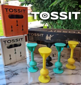 TOSSIT game expansion packs displayed on a marble surface, featuring colorful game pieces in yellow, green, and black, alongside packaging in orange and white, designed for interactive gameplay with options for 2 to 6 players.