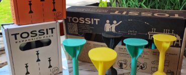 TOSSIT game expansion packs displayed on a marble surface, featuring colorful game pieces in yellow, green, and black, alongside packaging in orange and white, designed for interactive gameplay with options for 2 to 6 players.