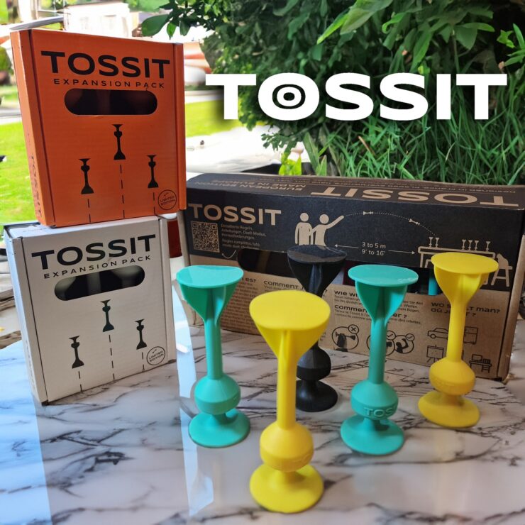 TOSSIT game expansion packs displayed on a marble surface, featuring colorful game pieces in yellow, green, and black, alongside packaging in orange and white, designed for interactive gameplay with options for 2 to 6 players.