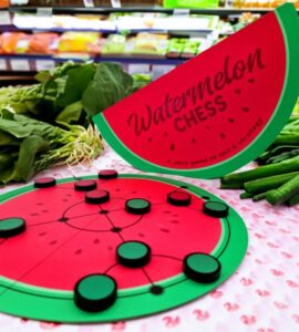 A colorful game of Watermelon Chess displayed among fresh vegetables, featuring a round watermelon-themed game board and a vibrant game piece design, showcasing a unique blend of fun and strategy in a grocery setting.