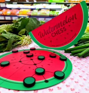 A colorful game of Watermelon Chess displayed among fresh vegetables, featuring a round watermelon-themed game board and a vibrant game piece design, showcasing a unique blend of fun and strategy in a grocery setting.