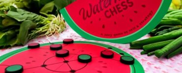 A colorful game of Watermelon Chess displayed among fresh vegetables, featuring a round watermelon-themed game board and a vibrant game piece design, showcasing a unique blend of fun and strategy in a grocery setting.