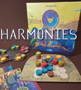 Colorful board game "Harmonies" by Johan Benvenuto featuring vibrant game pieces and a beautifully illustrated box. The game components include a hexagonal board, tokens in various colors, and cards laid out for gameplay, set against a rich brown background.