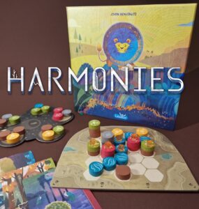 Colorful board game "Harmonies" by Johan Benvenuto featuring vibrant game pieces and a beautifully illustrated box. The game components include a hexagonal board, tokens in various colors, and cards laid out for gameplay, set against a rich brown background.