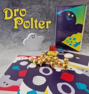 Image of the board game "Dro Polter," featuring a smiling ghost figure, colorful game cards with various designs, and game pieces including gold bells and a red block. The game box is visible, showcasing its vibrant artwork. Ideal for family game nights and fun gatherings.