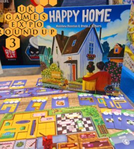 Image of the board game "Happy Home" by Matthew Dunstan and Brett J. Gilbert, displayed at the UK Games Expo. The game features vibrant artwork of a house and game components, including cards for different rooms like the kitchen and bathroom, all laid out on a wooden table. The background showcases the expo environment with colorful game displays.