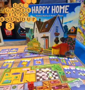 Image of the board game "Happy Home" by Matthew Dunstan and Brett J. Gilbert, displayed at the UK Games Expo. The game features vibrant artwork of a house and game components, including cards for different rooms like the kitchen and bathroom, all laid out on a wooden table. The background showcases the expo environment with colorful game displays.