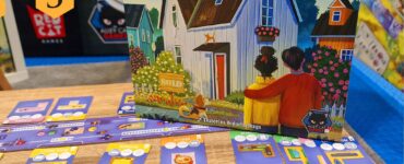 Image of the board game "Happy Home" by Matthew Dunstan and Brett J. Gilbert, displayed at the UK Games Expo. The game features vibrant artwork of a house and game components, including cards for different rooms like the kitchen and bathroom, all laid out on a wooden table. The background showcases the expo environment with colorful game displays.