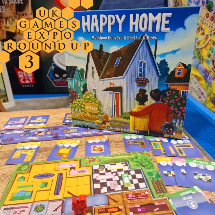 Image of the board game "Happy Home" by Matthew Dunstan and Brett J. Gilbert, displayed at the UK Games Expo. The game features vibrant artwork of a house and game components, including cards for different rooms like the kitchen and bathroom, all laid out on a wooden table. The background showcases the expo environment with colorful game displays.