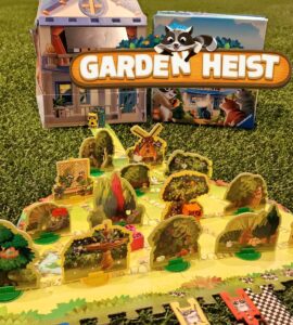 Colorful board game "Garden Heist" displayed on green grass, featuring a detailed game board with various garden elements, playful raccoon characters, and a charming house backdrop. Ideal for family game night and strategy enthusiasts.