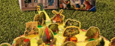 Colorful board game "Garden Heist" displayed on green grass, featuring a detailed game board with various garden elements, playful raccoon characters, and a charming house backdrop. Ideal for family game night and strategy enthusiasts.