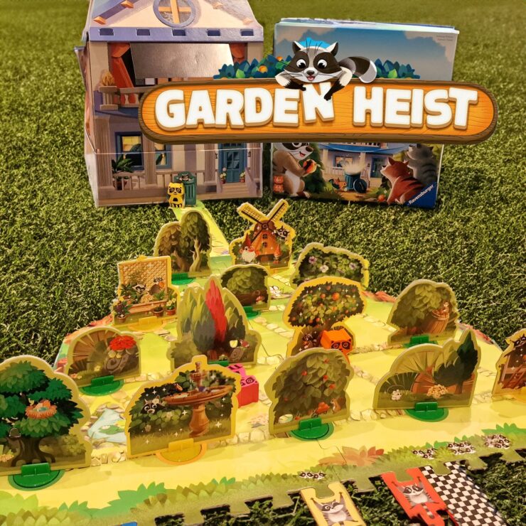 Colorful board game "Garden Heist" displayed on green grass, featuring a detailed game board with various garden elements, playful raccoon characters, and a charming house backdrop. Ideal for family game night and strategy enthusiasts.