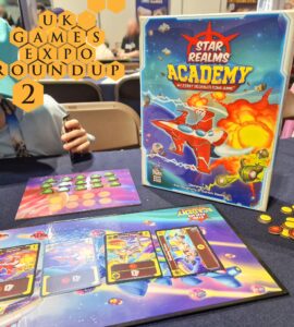 Image showcasing the board game "Star Realms: Academy" at the UK Games Expo, featuring the game box, player tokens, and game cards on a table. A child’s hand is visible holding a card, emphasizing the engaging and family-friendly nature of the game.