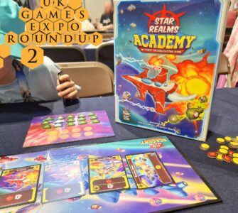 Image showcasing the board game "Star Realms: Academy" at the UK Games Expo, featuring the game box, player tokens, and game cards on a table. A child’s hand is visible holding a card, emphasizing the engaging and family-friendly nature of the game.