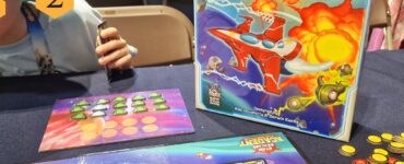 Image showcasing the board game "Star Realms: Academy" at the UK Games Expo, featuring the game box, player tokens, and game cards on a table. A child’s hand is visible holding a card, emphasizing the engaging and family-friendly nature of the game.