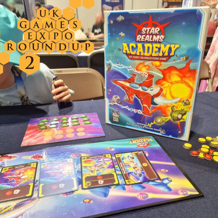 Image showcasing the board game "Star Realms: Academy" at the UK Games Expo, featuring the game box, player tokens, and game cards on a table. A child’s hand is visible holding a card, emphasizing the engaging and family-friendly nature of the game.