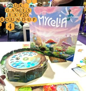 Image of the board game Mycelia displayed at the UK Games Expo, featuring the game box and a colorful game board with tokens. The scene captures a lively gaming environment with attendees in the background.
