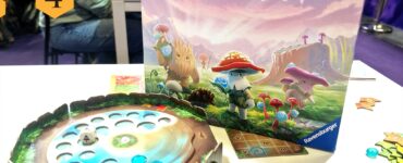 Image of the board game Mycelia displayed at the UK Games Expo, featuring the game box and a colorful game board with tokens. The scene captures a lively gaming environment with attendees in the background.