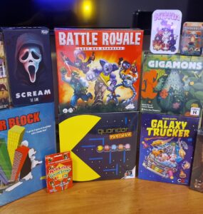 A collection of board games displayed on a wooden surface, featuring titles such as "Forever Home," "Scream: The Game," "Battle Royale," "Gigamons," "Tower Block," "Quoridor Pac-Man," "Galaxy Trucker," and "Paper Dungeons: Side Quest Expansion." The image showcases various game boxes with colorful artwork, appealing to board game enthusiasts.