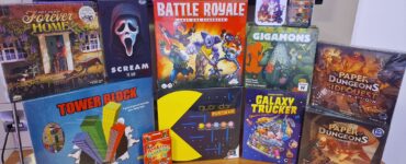 A collection of board games displayed on a wooden surface, featuring titles such as "Forever Home," "Scream: The Game," "Battle Royale," "Gigamons," "Tower Block," "Quoridor Pac-Man," "Galaxy Trucker," and "Paper Dungeons: Side Quest Expansion." The image showcases various game boxes with colorful artwork, appealing to board game enthusiasts.