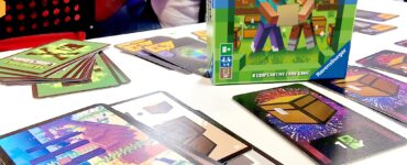 Image of the "Minecraft Explorers" cooperative card game by Ravensburger displayed at the UK Games Expo. The game box is prominently featured alongside various game cards spread out on a table, showcasing colorful graphics and gameplay elements. The background includes blurred figures, hinting at a lively expo atmosphere.