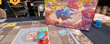 Board game "Robot Quest Arena" showcased at UK Games Expo Roundup 6, featuring colorful game pieces and a detailed game board. The image captures the vibrant box art alongside the gameplay in progress, with attendees visible in the background.