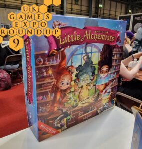 Colorful board game box for "Little Alchemists" displayed at the UK Games Expo, featuring whimsical characters engaged in potion-making. The box is labeled as a prototype and showcases vibrant artwork that captures the theme of alchemy and magic.