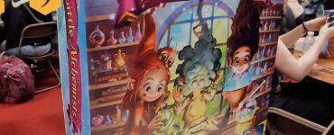 Colorful board game box for "Little Alchemists" displayed at the UK Games Expo, featuring whimsical characters engaged in potion-making. The box is labeled as a prototype and showcases vibrant artwork that captures the theme of alchemy and magic.