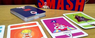 Anger Smash card game displayed on a wooden table, featuring the game box prominently and several colorful cards, including "Go Atomic" and "Yogamaster." The image highlights the game's vibrant artwork and engaging design, ideal for players seeking a fun, addictive card game experience.