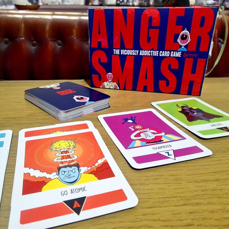 Anger Smash card game displayed on a wooden table, featuring the game box prominently and several colorful cards, including "Go Atomic" and "Yogamaster." The image highlights the game's vibrant artwork and engaging design, ideal for players seeking a fun, addictive card game experience.