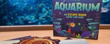 Colorful game box for "Escape from the Aquarium," an escape room game designed for kids aged 8-12, featuring underwater themes and playful graphics, displayed alongside game components on a table with an aquarium backdrop.