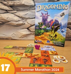 Colorful board game "Dragomino" displayed on a wooden table, featuring vibrant tiles and a pink dragon figure. The game box, illustrated with dragons and a fantasy landscape, is positioned against a textured stone backdrop. The image promotes the Summer Marathon 2024 event.