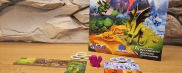 Colorful board game "Dragomino" displayed on a wooden table, featuring vibrant tiles and a pink dragon figure. The game box, illustrated with dragons and a fantasy landscape, is positioned against a textured stone backdrop. The image promotes the Summer Marathon 2024 event.