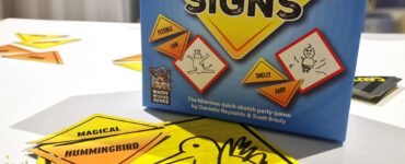 Caution Signs party game box displayed on a table with colorful sketch cards featuring humorous prompts. The image highlights the game's fun and interactive nature, suitable for gatherings and creative play. Summer Marathon 2024 branding is visible in the corner.