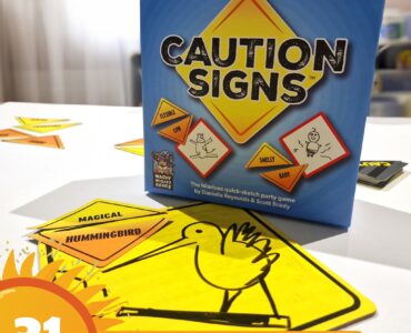 Caution Signs party game box displayed on a table with colorful sketch cards featuring humorous prompts. The image highlights the game's fun and interactive nature, suitable for gatherings and creative play. Summer Marathon 2024 branding is visible in the corner.