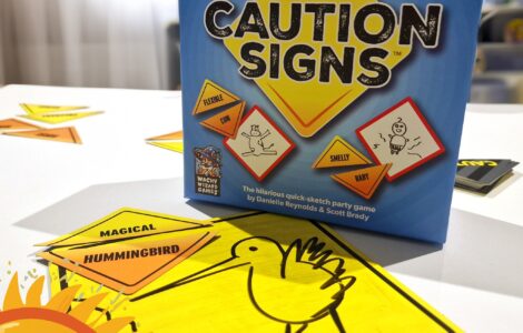 Caution Signs party game box displayed on a table with colorful sketch cards featuring humorous prompts. The image highlights the game's fun and interactive nature, suitable for gatherings and creative play. Summer Marathon 2024 branding is visible in the corner.
