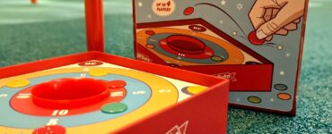 Colorful Tiddlywinks game by Super Retro Games, featuring a circular scoring board and a box design, suitable for up to four players. The image highlights the game setup on a green carpet, indicating its playful and engaging nature for family gatherings or game nights.
