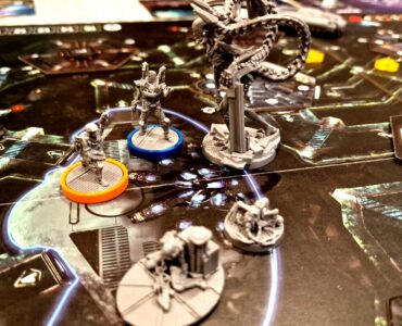 Close-up of a sci-fi board game in progress, featuring detailed miniatures of characters and creatures on a dark, intricate game board. The scene includes various player tokens in distinct colors and a central structure, highlighting the game's strategic elements and immersive artwork.