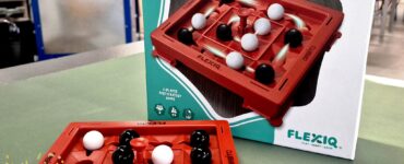 Image of the game "Orbito" by Flexiq, featuring a red game board with black and white game pieces arranged on it. The box is displayed in the background, highlighting its 2-player fast strategy gameplay. The image is part of the Summer Marathon 2024 event.
