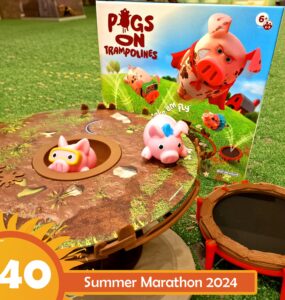 Colorful game setup of "Pigs on Trampolines," featuring two pig figures on a trampoline-like base, with the game box in the background. Ideal for children ages 6 and up, promoting fun and interactive play. Summer Marathon 2024 event branding visible in the corner.