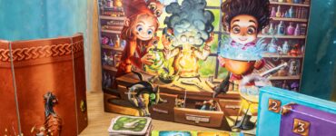 Colorful board game "Little Alchemists" displayed on a wooden table, featuring vibrant artwork of characters engaging in potion-making. The game box, cards, and tokens are arranged alongside a themed background, promoting the Summer Marathon 2024 event.
