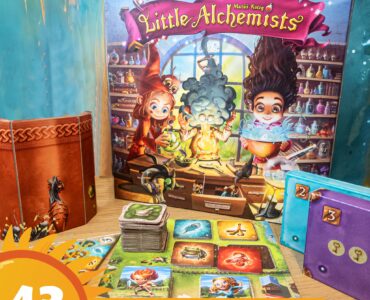 Colorful board game "Little Alchemists" displayed on a wooden table, featuring vibrant artwork of characters engaging in potion-making. The game box, cards, and tokens are arranged alongside a themed background, promoting the Summer Marathon 2024 event.