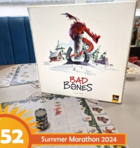 Board game "Bad Bones" by David Flies displayed on a table, featuring a colorful cover with a dragon and snowy landscape. Game pieces and cards are visible on the table, promoting the Summer Marathon 2024 event.