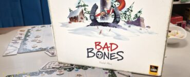 Board game "Bad Bones" by David Flies displayed on a table, featuring a colorful cover with a dragon and snowy landscape. Game pieces and cards are visible on the table, promoting the Summer Marathon 2024 event.