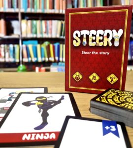 Board game "Steery" displayed with character cards, including a ninja, on a wooden table in front of a library backdrop. The game is suitable for 3-8 players, takes approximately 30 minutes to play, and is recommended for ages 10 and up.