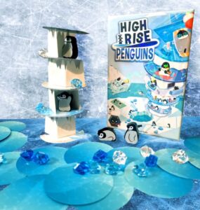 Colorful display of the board game "High Rise Penguins," featuring a multi-level penguin tower, game box, and decorative blue gems on a blue background. Ideal for family game nights and penguin enthusiasts.