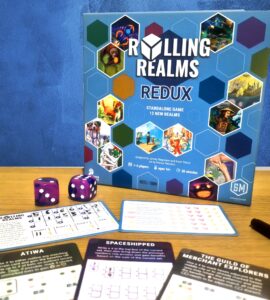 Board game "Rolling Realms: Redux" displayed with game components including dice and player sheets. The game is designed for 1-6 players, ages 14 and up, and features 12 new realms with a playtime of approximately 30 minutes.