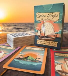 Spice Ships card game box and cards displayed on a wooden table at sunset. The box features a sailing ship design and a tagline about establishing trade routes. Nearby are several game cards, including "Nutmeg Island" and "Clove Ship," highlighting the game's theme of maritime exploration and trade.