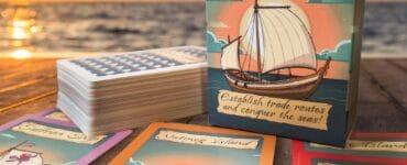 Spice Ships card game box and cards displayed on a wooden table at sunset. The box features a sailing ship design and a tagline about establishing trade routes. Nearby are several game cards, including "Nutmeg Island" and "Clove Ship," highlighting the game's theme of maritime exploration and trade.